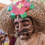 Colorful mask dancer wearing a straw hat with bright flowers during a traditional festival parade, showing mexico holiday season celebrations and local folklore.